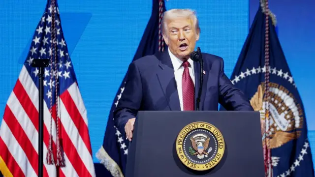 U.S. President Donald Trump delivers remarks at the America Business Forum in Miami, wearing a blue suit and red tie. He speaks into a microphone with flags behind him.