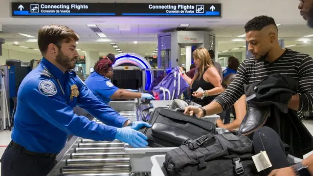 A TSA agent in blue shit, blue gloves and short brown hair reaches over to pick up the bag of a passenger going through security.