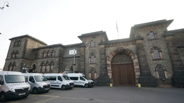 Wandsworth prison taken from in front of the main entrance, showiing a victorian sont building with large arched wooden door. There are four silver prison service vans parked outside