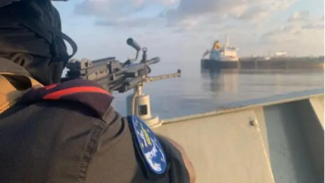 A sailor with a EUNavfor sleeve patch trains a machine gun from a navy vessel on an oil tanker in the distance