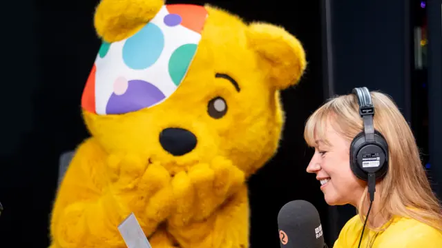 Sara Cox wearing headphones alongside a life-size Pudsey Bear
