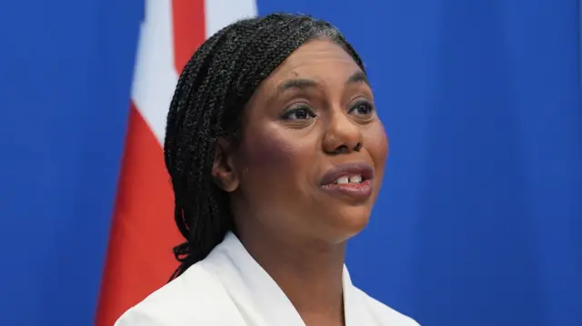 Close crop of Badenoch from shoulders up. Blue background and partial union jack. She is wearing white