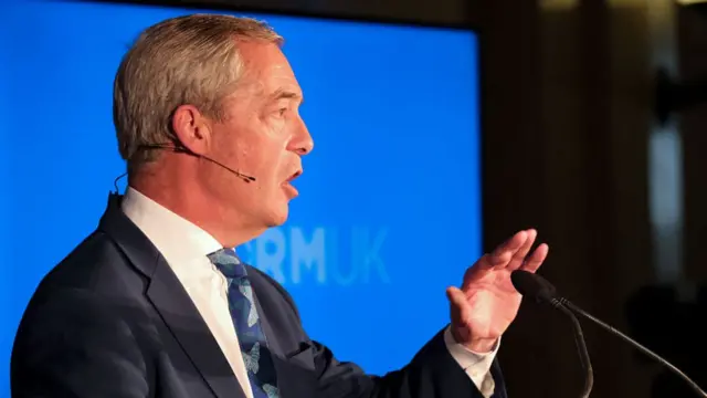 Nigel Farage in a suit talking into a microphone, his hand is outstretched and there is a blue and black background.