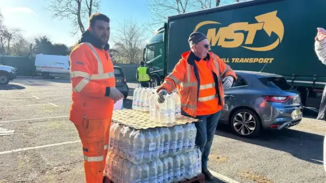 Two men in hi vis jackets with piles of water bottles