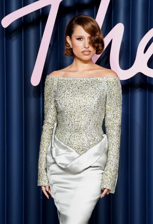 Singer Raye wears a silver and gold glittering top with a shining, draped skirt below. She stands in front of a blue background.