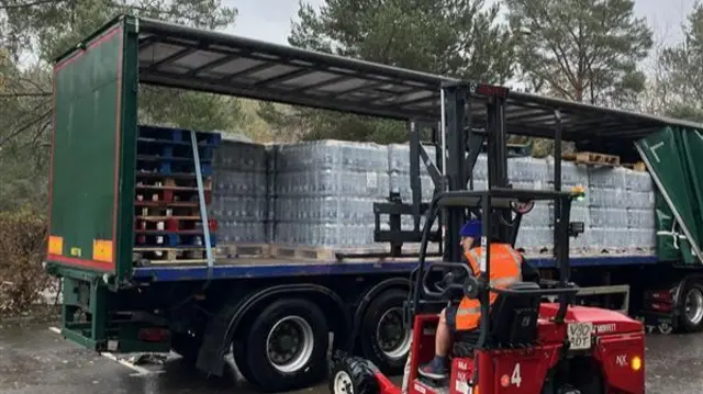 A man in an orange high-vis jacket driving a small, red forklift. The vehicle's forks are inserted into a pallet on the back of a curtain trailer. The pallet is loaded with stacks of large water bottles, and is one of several such loads seen behind the open trailer.