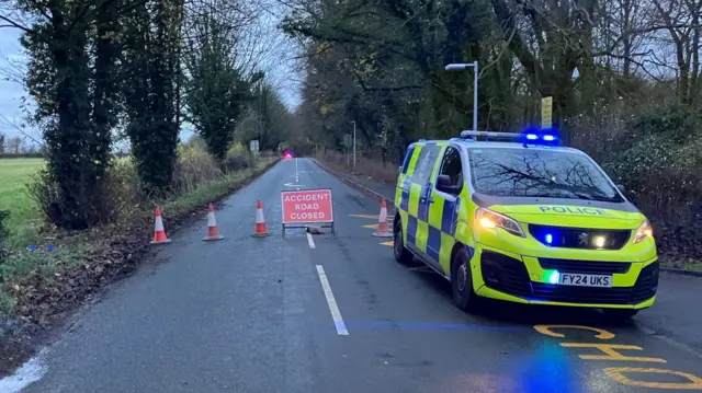 Rural road closed due to a crash, with a police car displaying flashing blue lights and high-visibility markings parked beside traffic cones and a red "Accident Road Closed" sign; surrounding landscape includes bare trees and open fields under an overcast sky.