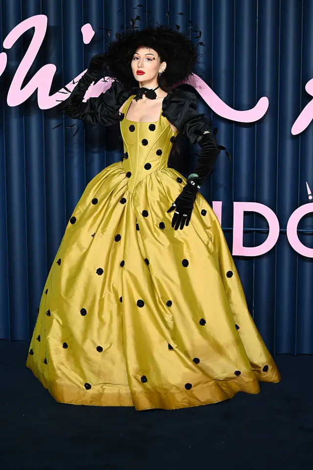 Eve Lily Parsley wears a yellow dress with black spots and a dramatic feathered, wide-brimmed hat