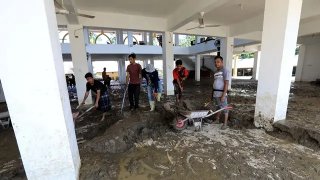 People holding wheelbarrows and sticks trying to clear mud from the inside of a large building