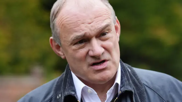 Close up of Ed Davey speaking to the press, he is wearing a grey jacket and white shirt and looking off camera