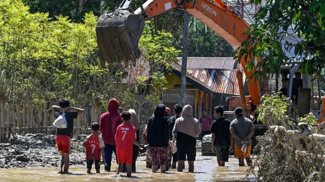 Backview of women and children wading in knee-deep brown water on a road. there is an excavator in front of them