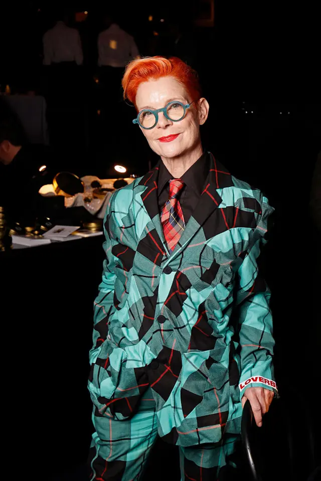 Sandy Powell wearing a green, black and red checked suit and turquoise-framed glasses