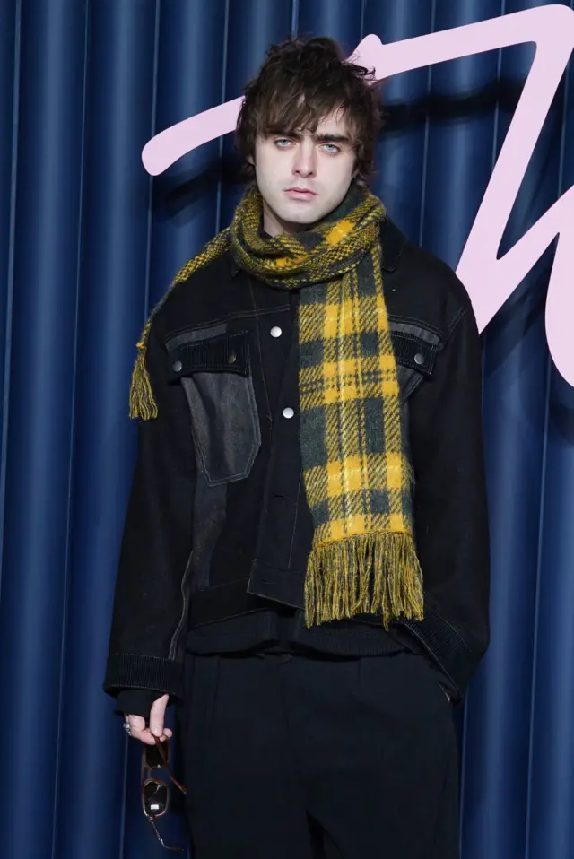 Model Lennon Gallagher wears a yellow and dark-blue tartan scarf over black clothing.