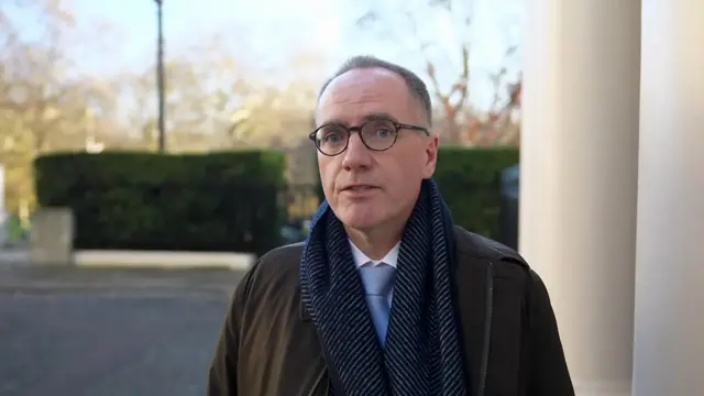 Former OBR chair Richard Hughes stands outdoors in a dark coat, blue scarf and light blue tie mid-speech. Two columns and some green shrubbery behind him