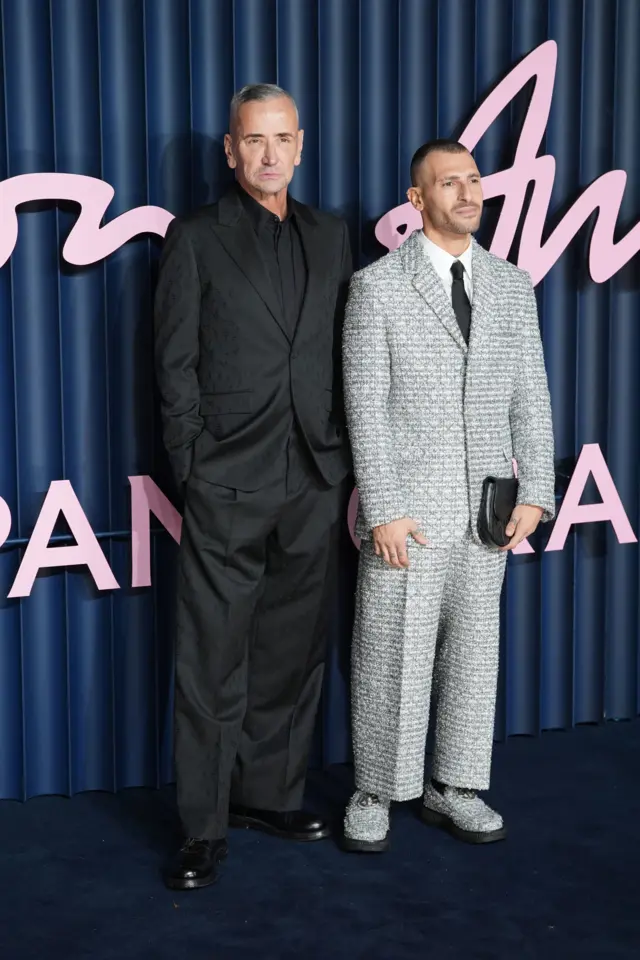 DJ Fat Tony wears an all-black suit and his husband Stavros Agapiou wears a grey, textured suit