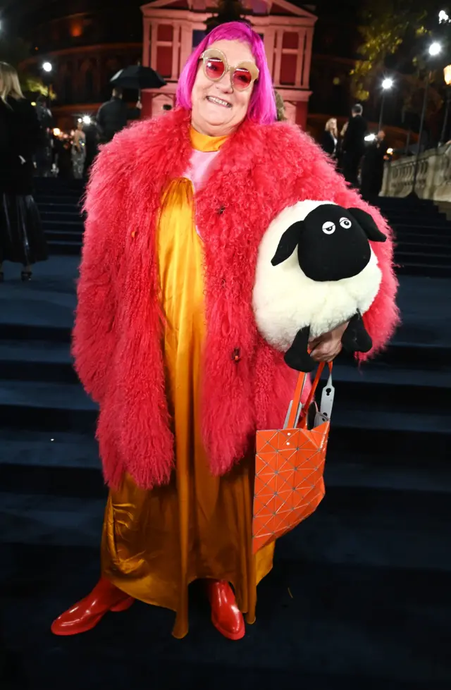 Berni Yates wears in a yellow dress, shaggy pink coat and a stuffed sheep under their arm