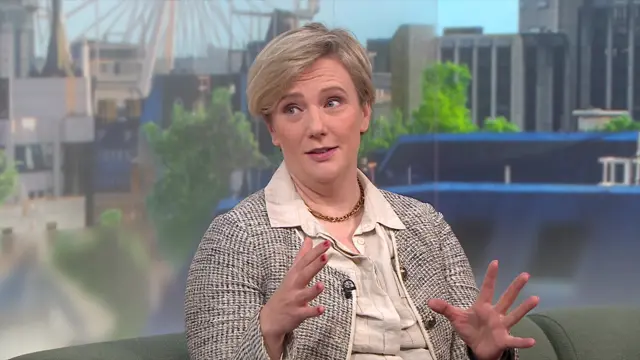 Stella Creasy sitting down on a green couch wearing a black and beige blazer and beige shirt, her hands raised as she gesticulates mid-speech