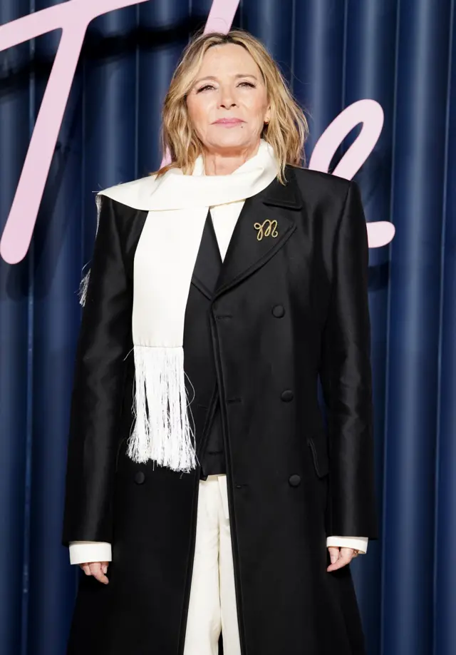 Actress Kim Cattrall wears a shining, black double-breasted coat with a white scarf draped over her front.