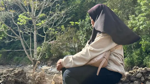 A woman in a beige shirt, black pants and black headscarf  sits in a mound of dirt and debris, looking sideways to the camera