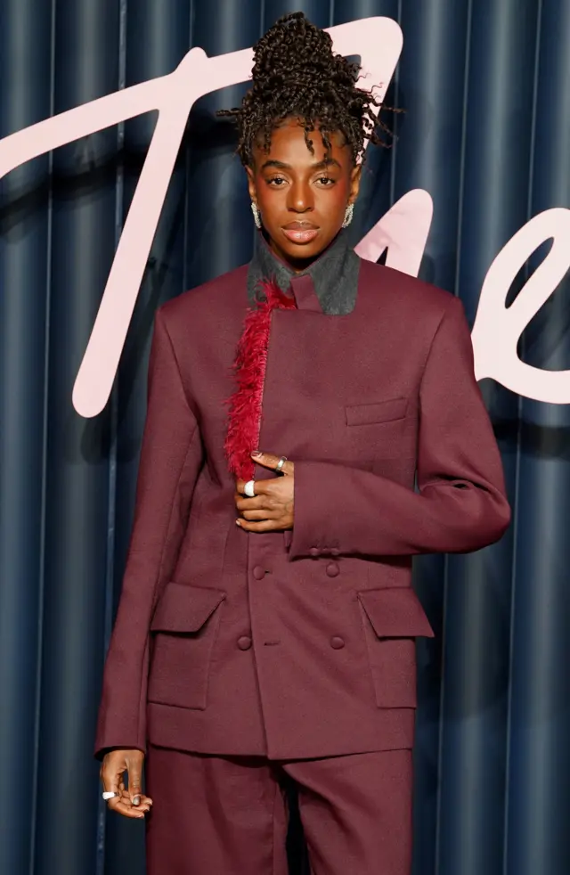 Singer Cat Burns wears a maroon trouser suit with a bit of red feathering on the jacket.