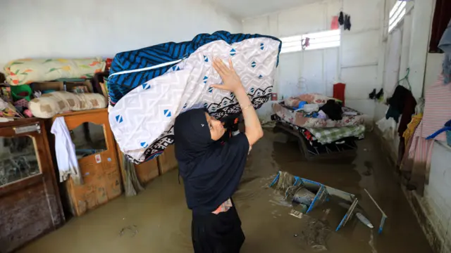 A young woman in a black tudung carrying a large cloth box on her shoulder. She is knee-deep in water in a home surrounded by damaged furniture