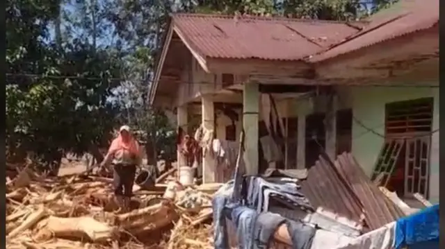 A woman walking among branches strewn all over the ground, in front of a one-storey house