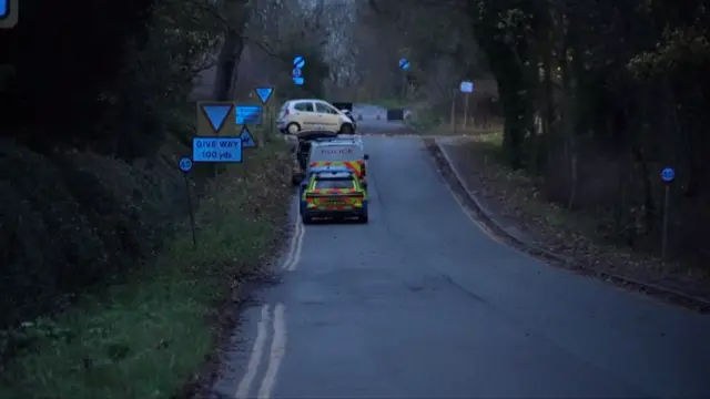 Two police vehicles parked along a country lane with a light-coloured car at the end and a cordon in the distance.