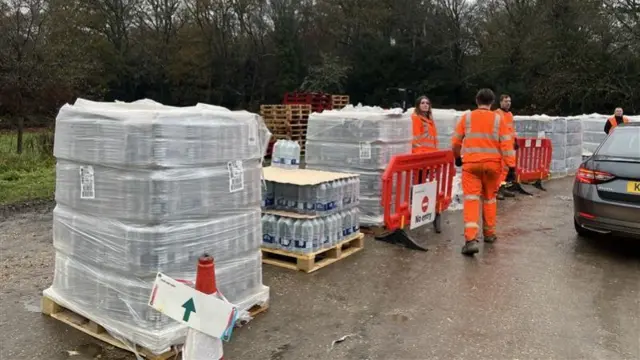 Rows of pallets containing cases of bottled water in a row. A car is parked beside them and there are three people wearing high-vis clothes attending them.