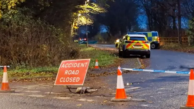 Road closed due to accident, marked by a red "Accident Road Closed" sign, orange traffic cones, and blue-and-white police tape; two police vehicles with flashing lights are parked in the background, with a "No Entry" sign to the left and fallen leaves scattered along a tree-lined road under dim lighting.