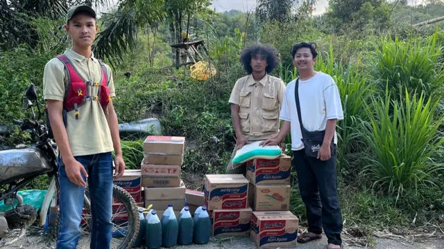 Three men posing for a photo besides boxes and bottles of relief items, like instant noodles