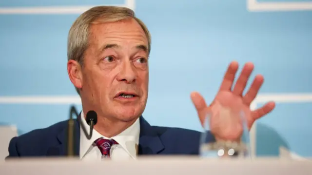 Nigel Farage in blue suit and patterned tie mid-speech, his left hand extended in front of him. He appears to be sitting behind a table, a thin black mic in front of him