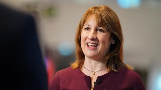 Rachel Reeves speaks to a journalist out of shot. She wears a maroon dress and gold necklace