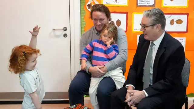 A child with red hair holds her hand above her head, as a father sits with his child on his lap and Starmer sits to the right