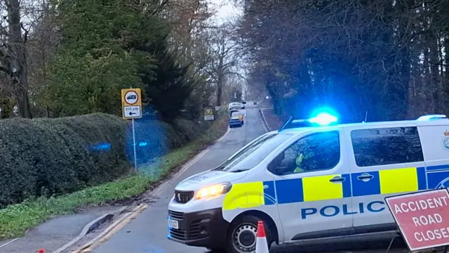 Police van with flashing blue lights blocks a rural road following an accident; a red "Accident Road Closed" sign and traffic cones mark the closure.