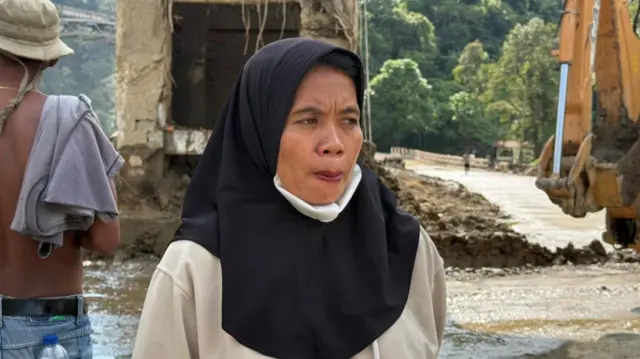 A woman in a black headscarf stands in front of a river, a ruined bridge and an excavator