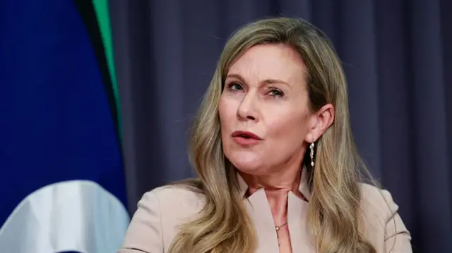 A smartly dressed woman with long blonde hair speaks against a background of blue curtains.