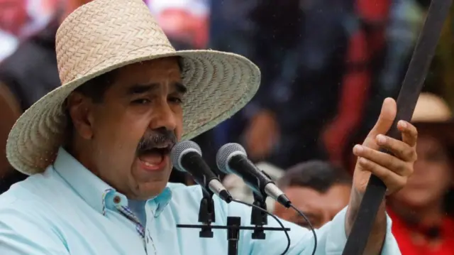 Nicolas Maduro in a light blue shirt and straw hat holds up a wooden stick whilst speaking into two mics. There's a large group of people behind him in the blurred background