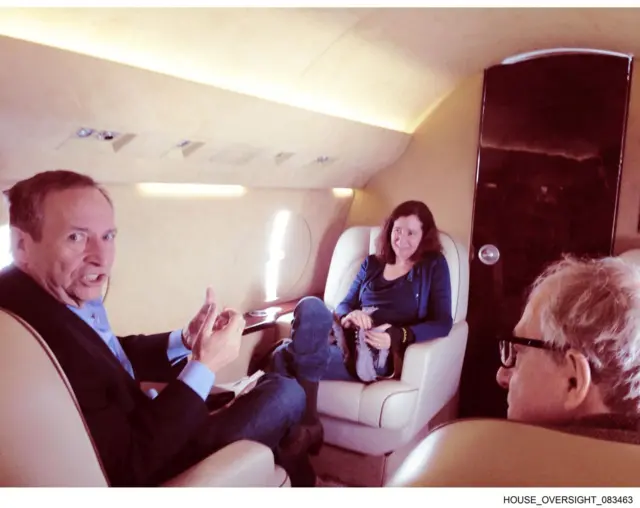 A photo of Larry Summers inside a jet with cream coloured walls. He can be seen looking at the camera with his hands raised in front of him.