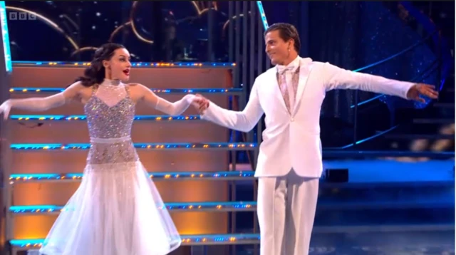 Amber and Nikita in matching white gown and suit holding hands during performance