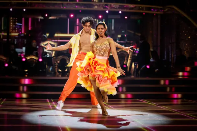 Julian (L) and Balvinder (R) in yellow and orange clothing dancing to salsa