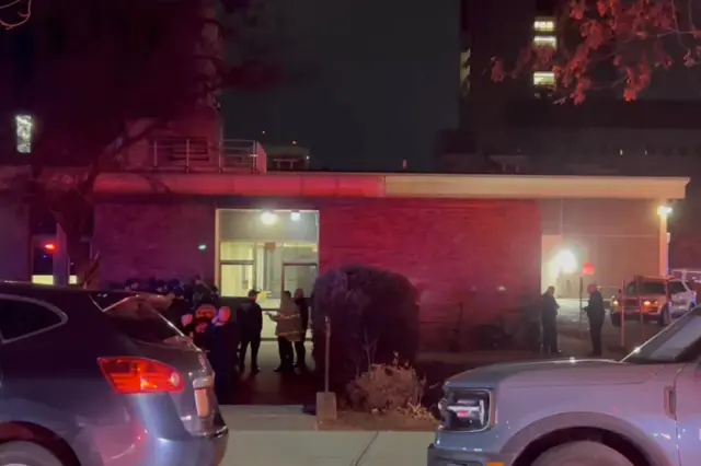 Fire fighters and police officers outside a brick building, red sirens reflected on the walls