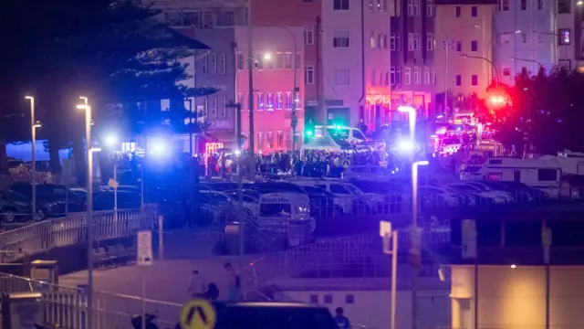 Wide shot of a car park near Bondi Beach with multiple emergency respondents cars with sirens on following a shooting
