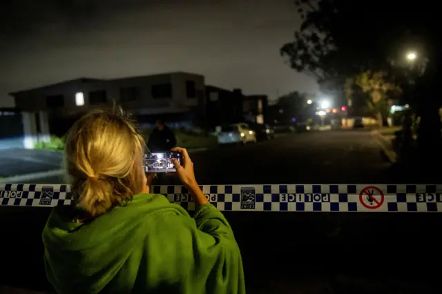 A woman records on her mobile phone as a police crime scene is established at the home of a suspect in Bonnyrigg,