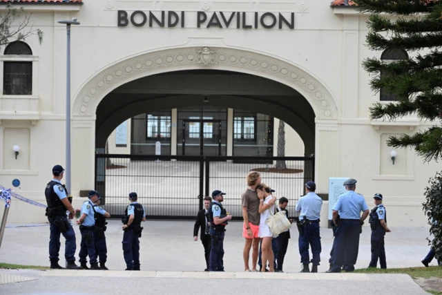 Two people embrace as police officers stand guard outside Bondi Pavilion as officers stand guard