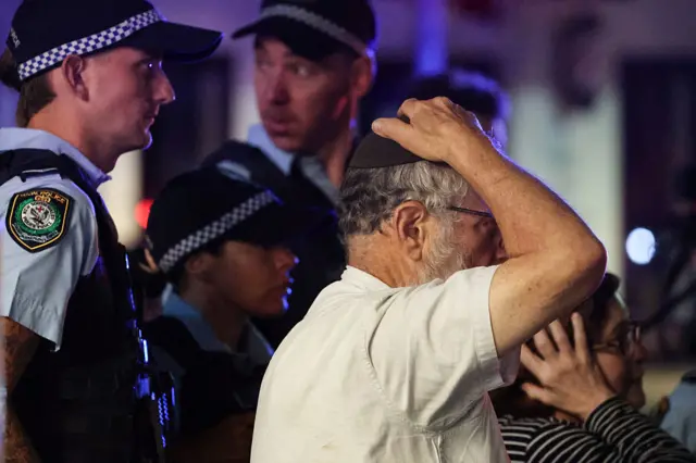 Police officers stand at the scene, a man in a white shirt and vest has his right hand on top of his head, holding his dark kippah