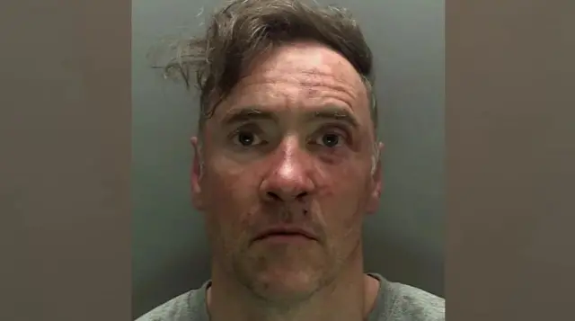 Custody mugshot of Paul Doyle, who has untidy greying brown hair swept over to one side. He has a neutral facial expression and is wearing a grey t-shirt.