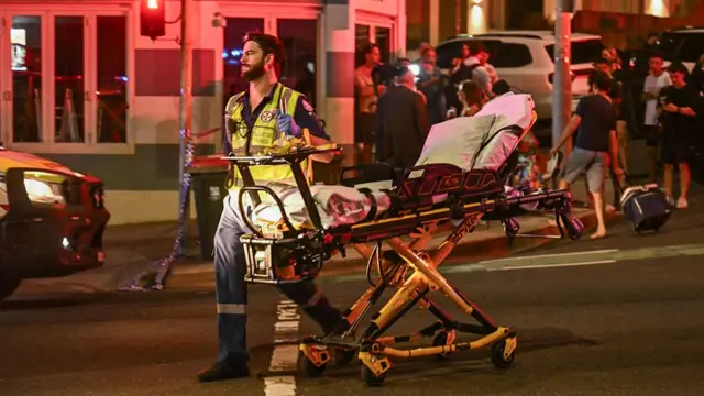 A first responder pushes an empty gurney across a street, several people milling around a building behind him