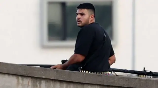 A gunman stands on a bridge wearing a black t-shirt. He is holding a weapon with ammunition around his waist. A gun and other ammunition appears next to him on the bridge