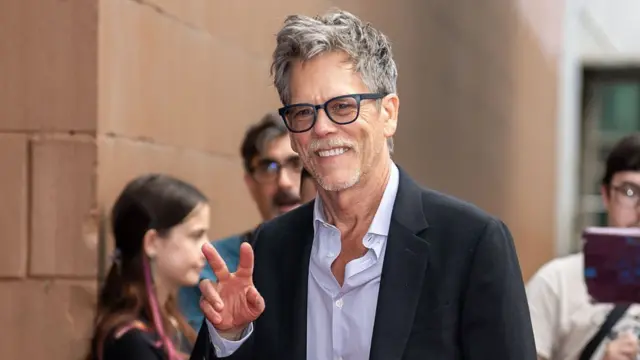 Kevin Bacon wears a black jacket and light coloured shirt as he walks outside. He smiles and holds up a peace sign