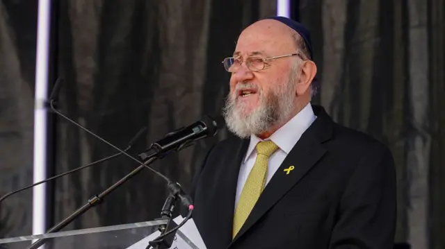 Sir Ephraim Mirvis speaking into a microphone, wearing a black suit and yellow tie